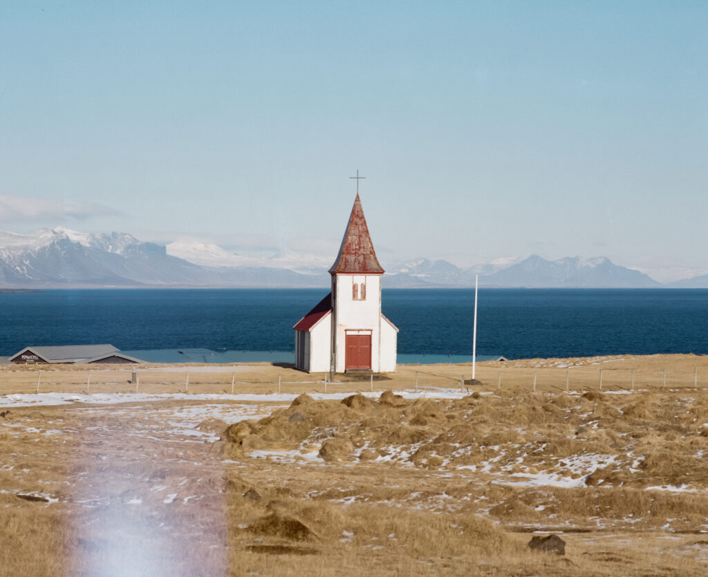 Photo d'une église prise en Islande. En arrière plan il y a la mer et au premier plan, de l'herbe desséché.